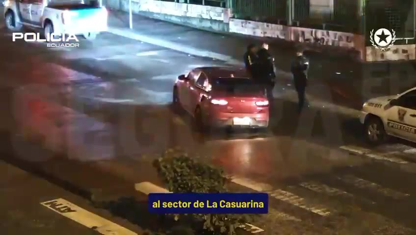 Imagen nocturna de videovigilancia: un auto rojo detenido en una calle con policías alrededor.
